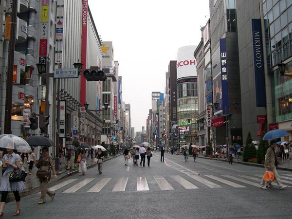 Pedestrians with umbrellas cross the wet street in Ginza among bright storefronts