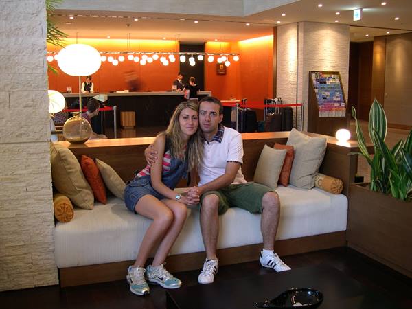Couple sitting on white sofa in hotel lobby, lit reception desk behind them