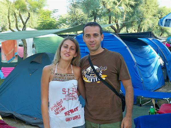 Smiling couple in front of blue camping tents, trees and shade tarps above
