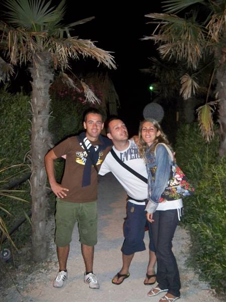 Three friends smile under palm trees at night, wearing flip-flops