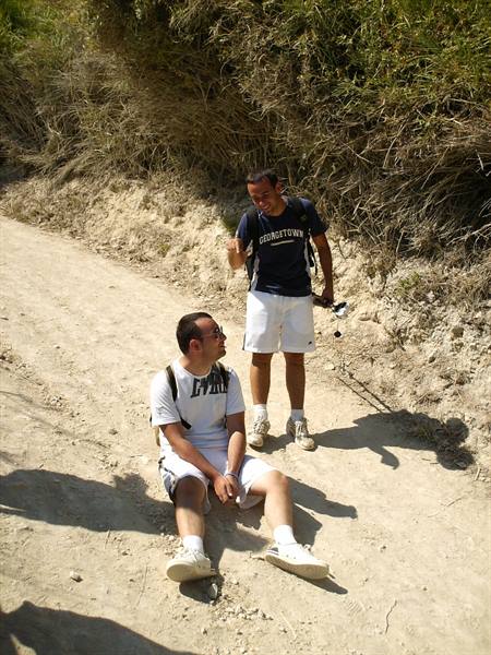 Two hikers resting on a dirt road, one sitting and one standing