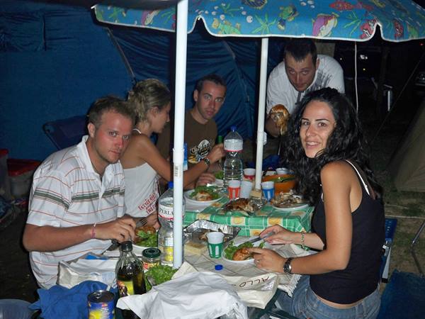 Friends dining under a patio umbrella at a campsite, tents behind, bottles on table