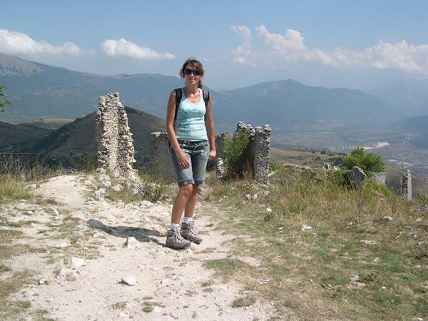 Girl with backpack standing among old stone wall ruins, mountains behind
