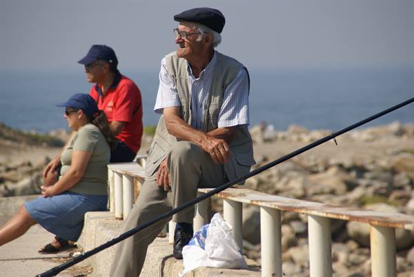 Elderly man in beret with fishing rod sitting on a low sea wall