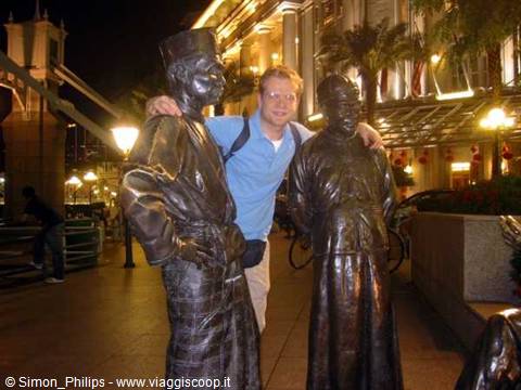 Tourist posing between two bronze statues at night by Singapore river