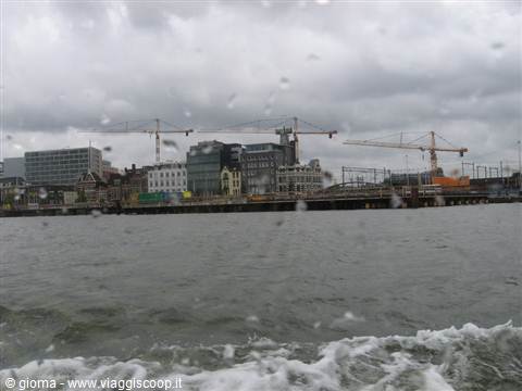 Cranes and buildings along the waterfront seen from a ferry under grey sky