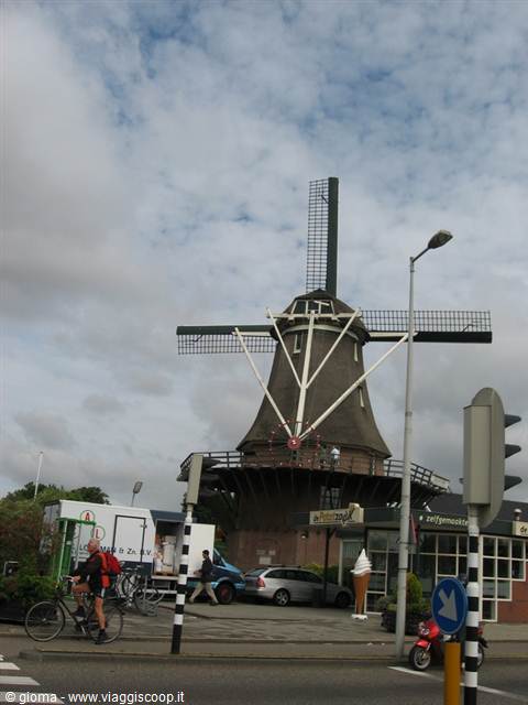 Brick windmill at a city crossroads, cyclist and cars under cloudy sky