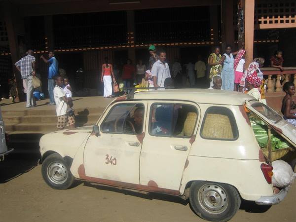 Old rusty white Renault 4 in front of a busy market, people on steps