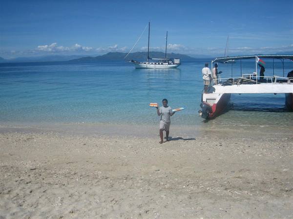 Man carries trays on sandy beach, catamaran and sailboat anchored