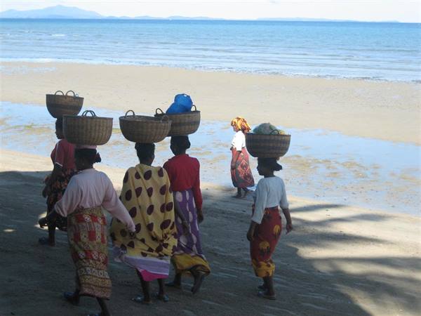 Women carrying woven baskets on their heads walk along the sunny beach