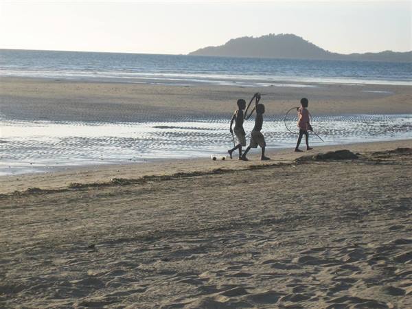 Children walk with fishing nets on a sandy beach by a hilly island