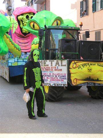 Carnival parade: person in green skeleton costume beside an allegorical float pulled by a tractor