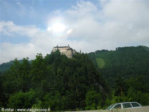 Stone castle on a forested hill, cloudy sky, car in the foreground