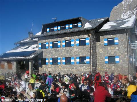 Stone mountain hut with blue shutters crowded with skiers in the sun