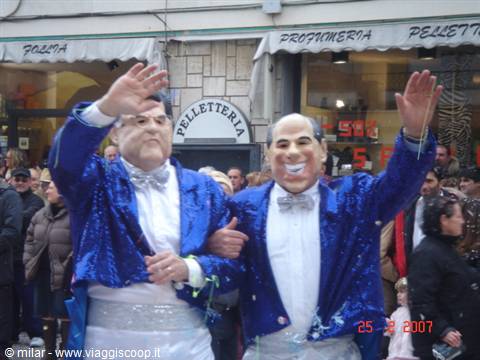 Two grinning masked men waving in blue jackets before a shop crowd
