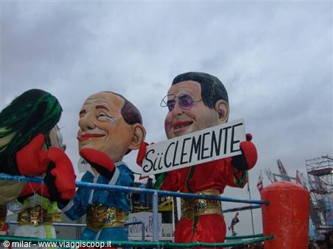 Carnival float with papier-mâché political figures, one puppet holding a Sù Clemente sign