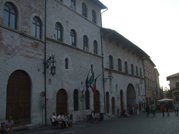 White stone palace with Italian flags, people sitting on the square in Assisi
