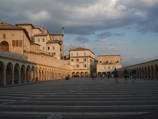 Assisi, Lower Piazza San Francesco: stone arcades and tourists in evening light