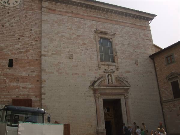 Light stone church facade with carved portal, visitors entering, Spello