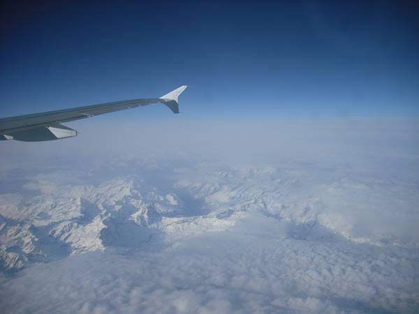Airplane wing above snowy Alps emerging from clouds, clear blue sky