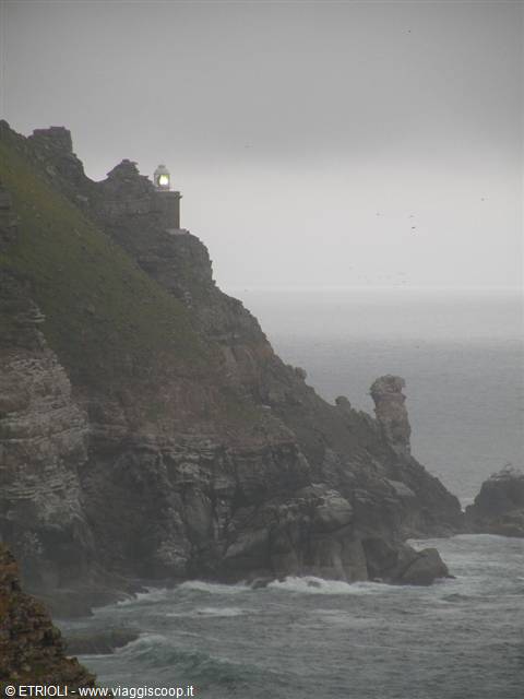 Lit lighthouse on rocky Cornish cliffs under grey sky, waves below
