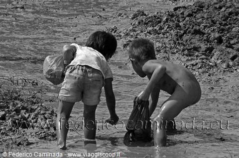 Two children collecting things in mud with plastic bags, harsh light