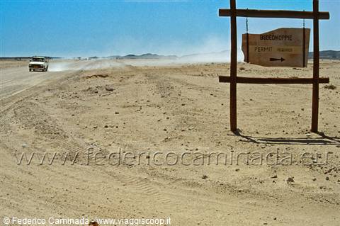 Wooden sign reading Sossusvlei on sandy desert track, car raising dust