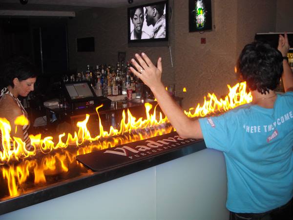 Waiter raises arms beside flames along the bar counter, TV above