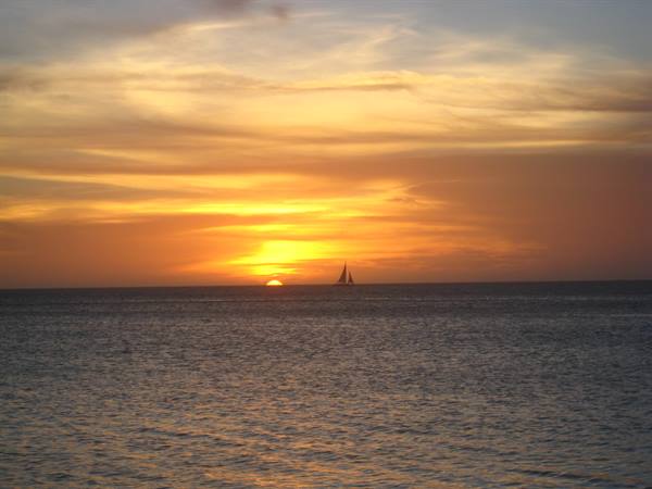 A sailboat crosses the golden sea beneath the setting sun
