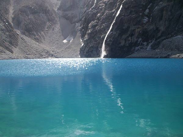 Turquoise lagoon below grey rock walls, white waterfall and sunlight sparkling on water