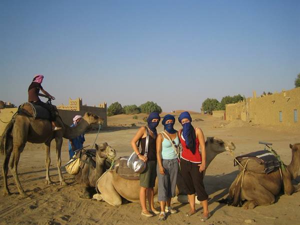 Three tourists in blue headscarves beside resting camels near mud-brick houses
