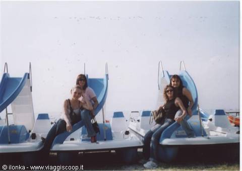 Four girls sitting on white and blue pedal boats with slides, sunny shore