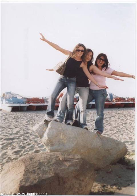 Three friends in jeans on a beach rock, arms outstretched, boats on sand