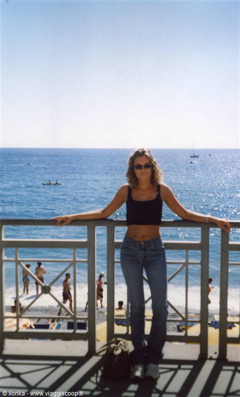 Girl leaning on railing above beach, blue sea with boats and swimmers