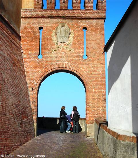 Tourists under the brick castle gate adorned with a stone coat of arms