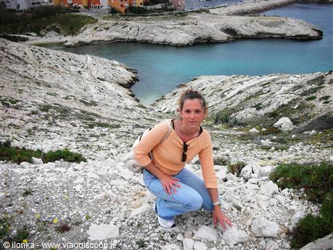 Woman in orange sweater kneeling on white rocks above a turquoise cove