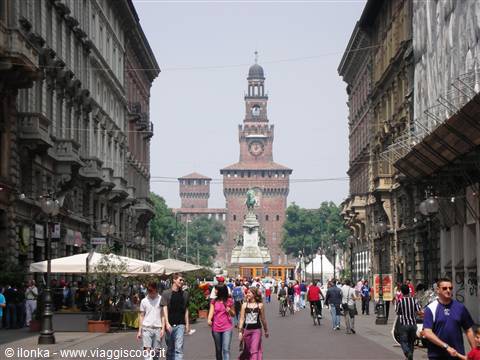 Crowded Via Dante toward Milan's Castello Sforzesco, brick crenellated tower