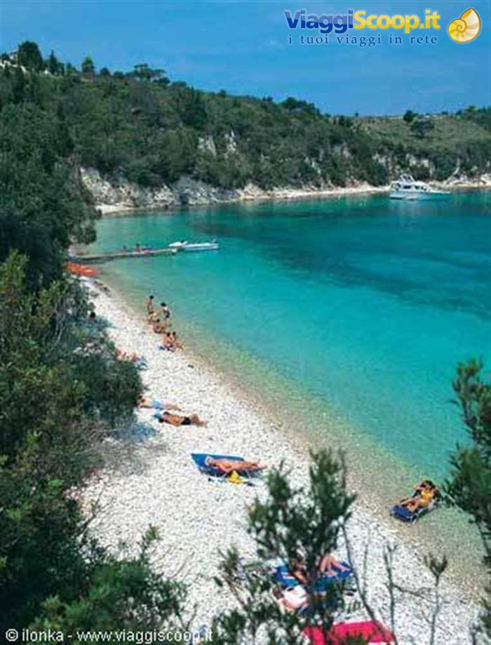 Beachgoers on pebble beach, turquoise water, boats and green trees
