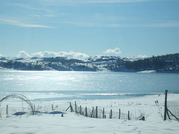 Frozen lake among snowy hills, wooden fence on snow, bright light