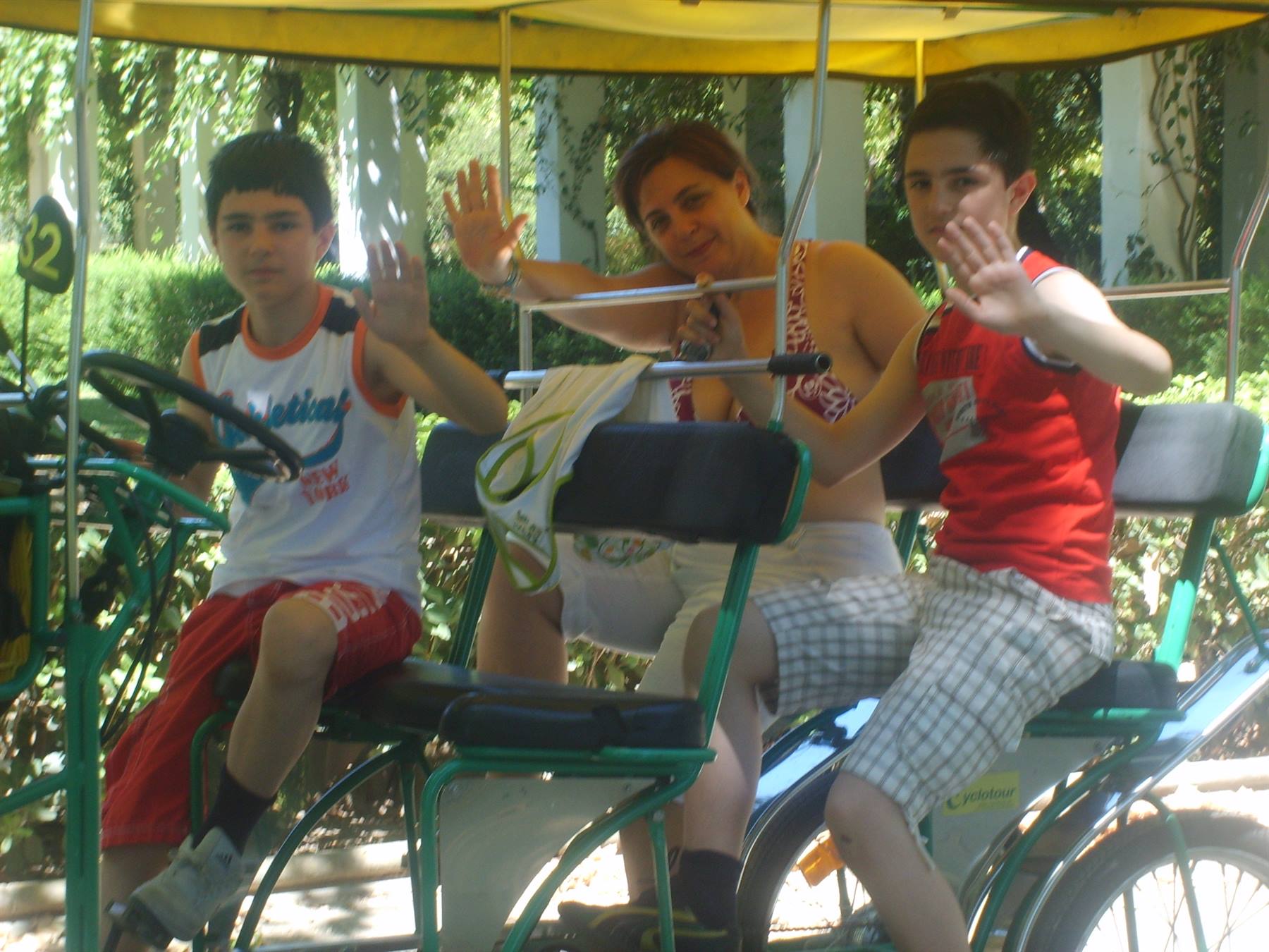 Woman and two boys waving from a pedal cart in a sunny park