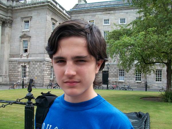 Boy in blue t-shirt in front of a grey stone building, bicycles parked on the lawn