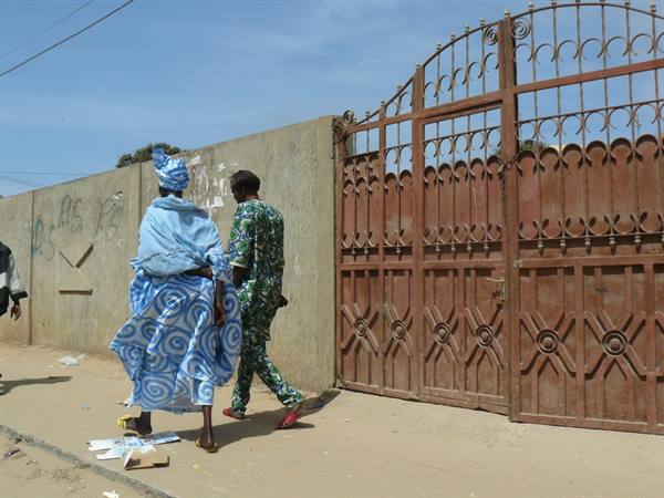 Two men in colorful boubous walk past a large rusty iron gate