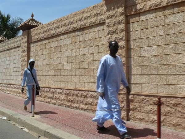 Two men in light blue boubous walk past a stone wall