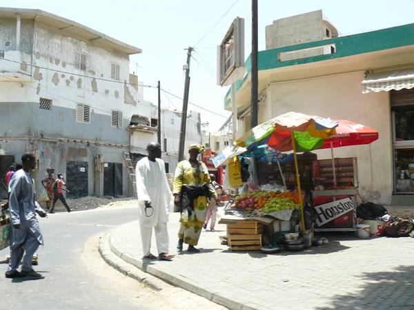 Fruit vendor under colorful umbrellas at a street corner, men in boubous passing by