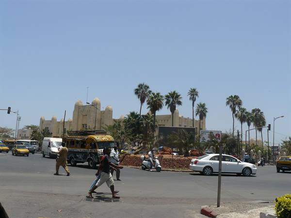 Pedestrians and cars crossing a roundabout with palms and arched building