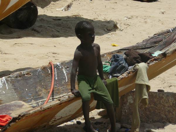 Child in green pants sitting on a wooden pirogue by the sandy beach