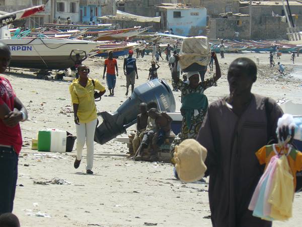 Fishermen and colorful boats on sandy beach, man selling clothes, woman carrying basket on head