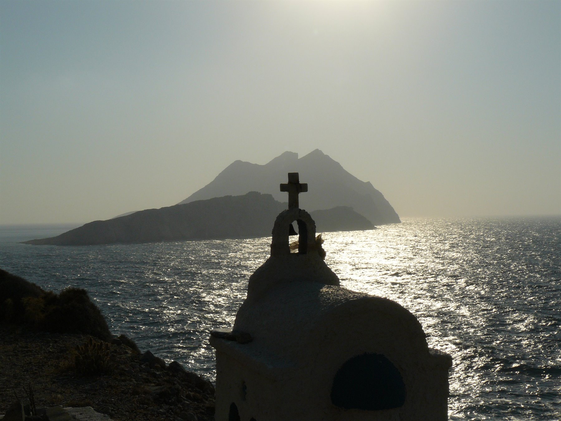 Campanile bianco con croce su mare lucente, isola vulcanica sullo sfondo