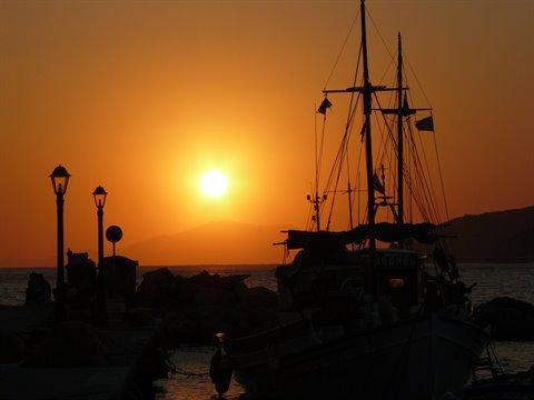 Sailing boat silhouetted at the pier with lampposts, orange sun over the sea