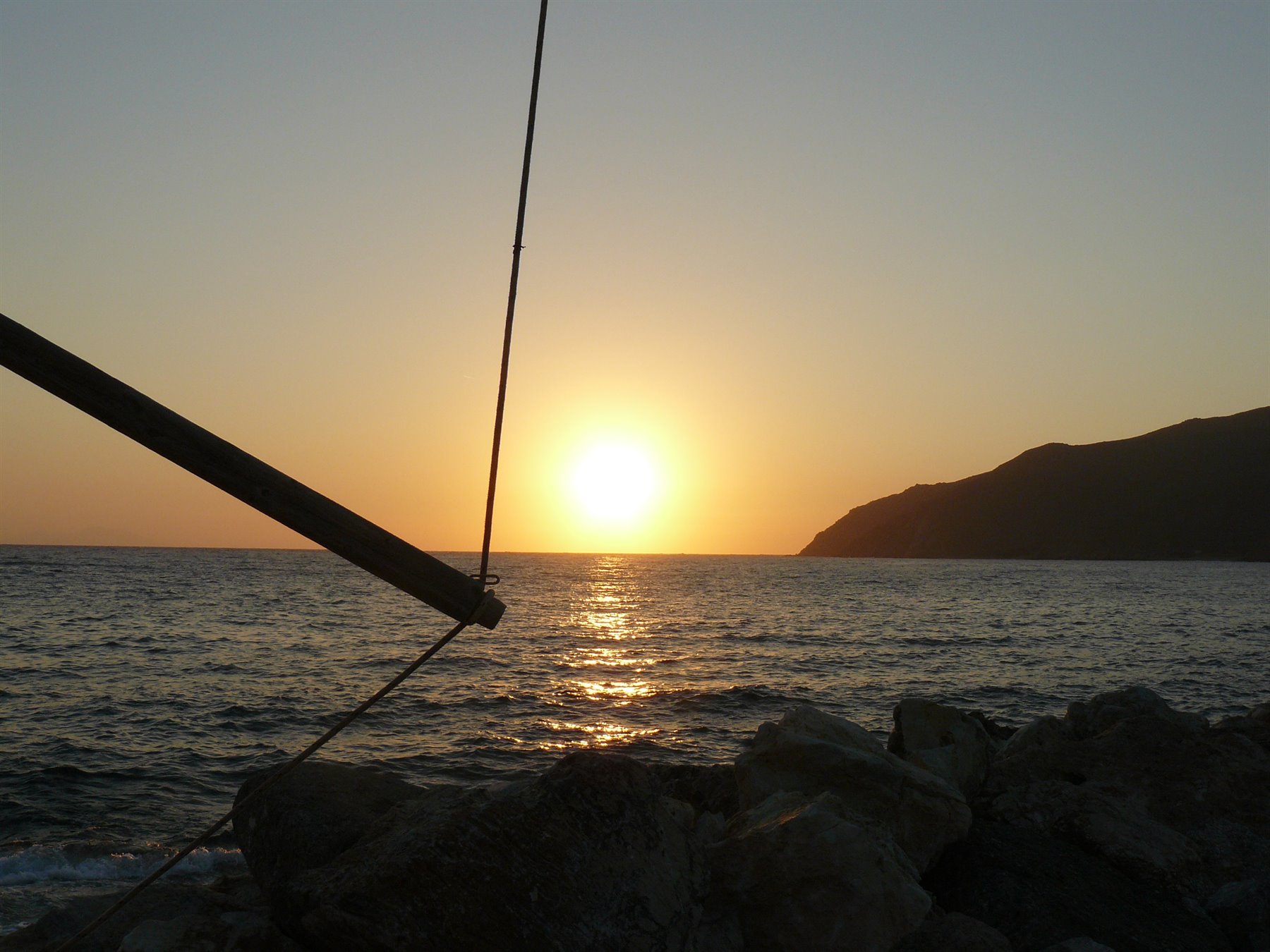 Sole basso sul mare davanti a scogli e una montagna scura, con un albero di barca in primo piano